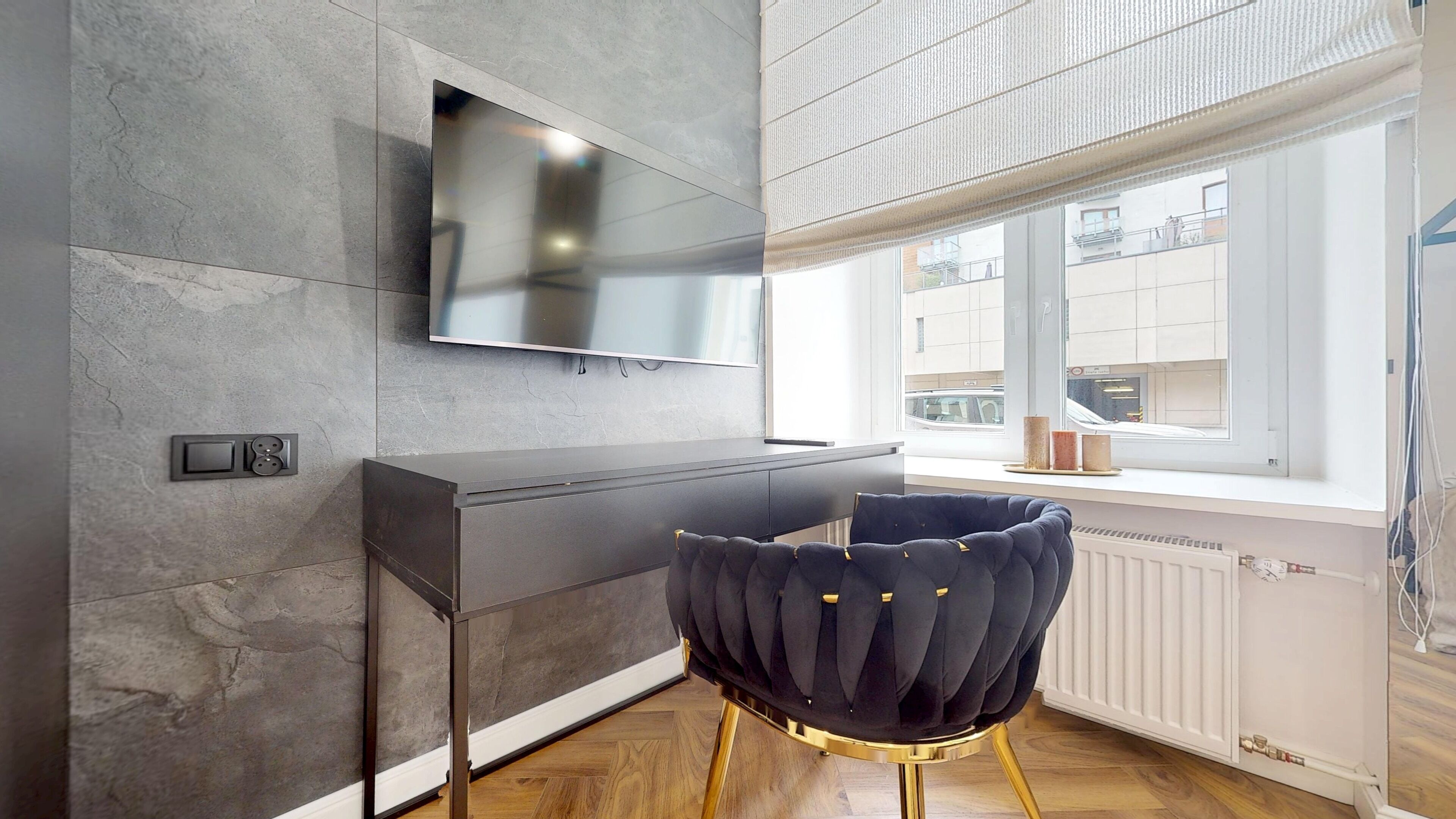 A modern living room with a flat-screen TV mounted on the wall, a dark sofa, and a window with a view of the city.