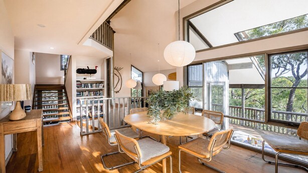 A spacious room with a wooden dining table, chairs, and a large window offering a view of trees. - Coastal Wilderness Retreat (HARDYS BAY)