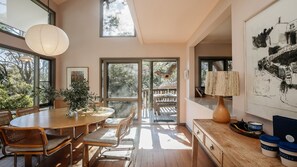 A dining area with a wooden table and chairs, a hanging light fixture, and a view through glass doors to an outdoor deck. - Coastal Wilderness Retreat (HARDYS BAY)