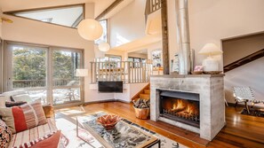 A spacious living room with a fireplace, wooden flooring, and a large window providing a view of the outdoors. - Coastal Wilderness Retreat (HARDYS BAY)