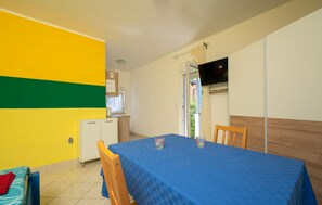 Ein Zimmer mit einem blauen Tisch, zwei Stühlen, einer kleinen Küchenzeile, einem Fernseher und einem Fenster mit Vorhängen.
