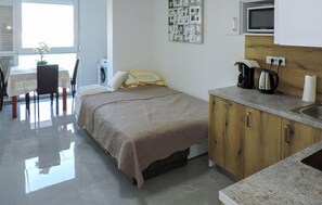 Ein kleines Schlafzimmer mit Bett, einer kompakten Küchenzeile, einem Essbereich und einem Fenster mit Jalousien.