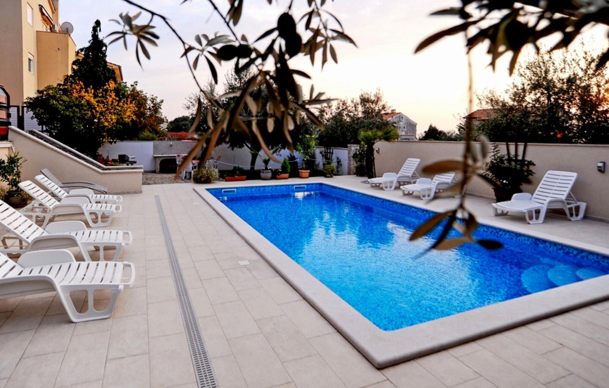 Une piscine rectangulaire entourée de chaises longues blanches, avec des plantes en pot tout autour et un bâtiment visible à l’arrière-plan.