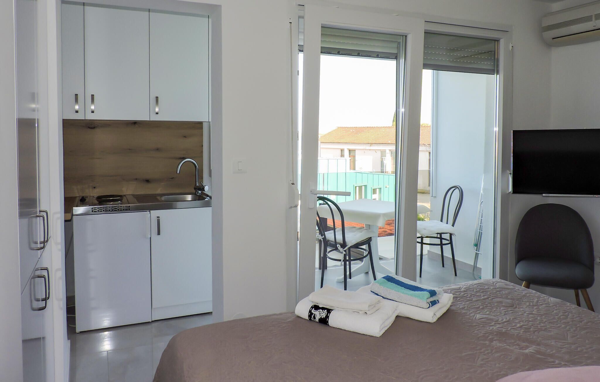 A hotel room with a bed, a desk, a chair, a TV, a kitchenette, and a sliding glass door to the outside.