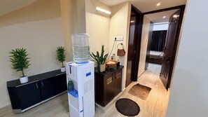 Uma área de recepção com bebedouro, plantas em vasos, uma pequena mesa e uma porta que dá para um quarto.