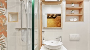Un baño moderno con cabina de ducha de vidrio, un lavamanos blanco y un estante de madera con artículos de aseo.