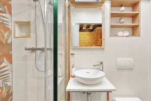 A modern bathroom with a glass shower enclosure, a white sink, and a wooden shelf with toiletries. - Cosy Studio Near Louvre & Les Halles (Paris)
