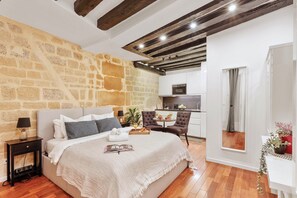 A bedroom with a stone wall, a bed with a white bedspread, a bedside table with a lamp, and a view into the kitchen. - Cosy Studio Near Louvre & Les Halles (Paris)