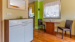 Et lille værelse med tekøkken, spiseplads med bord og stole, et vindue med let gardin og et spejl på væggen.