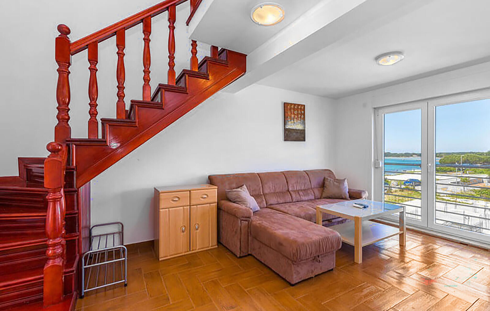 Sala de estar com escada de madeira, um sofá, uma mesa pequena e vista para o mar através de grandes janelas.