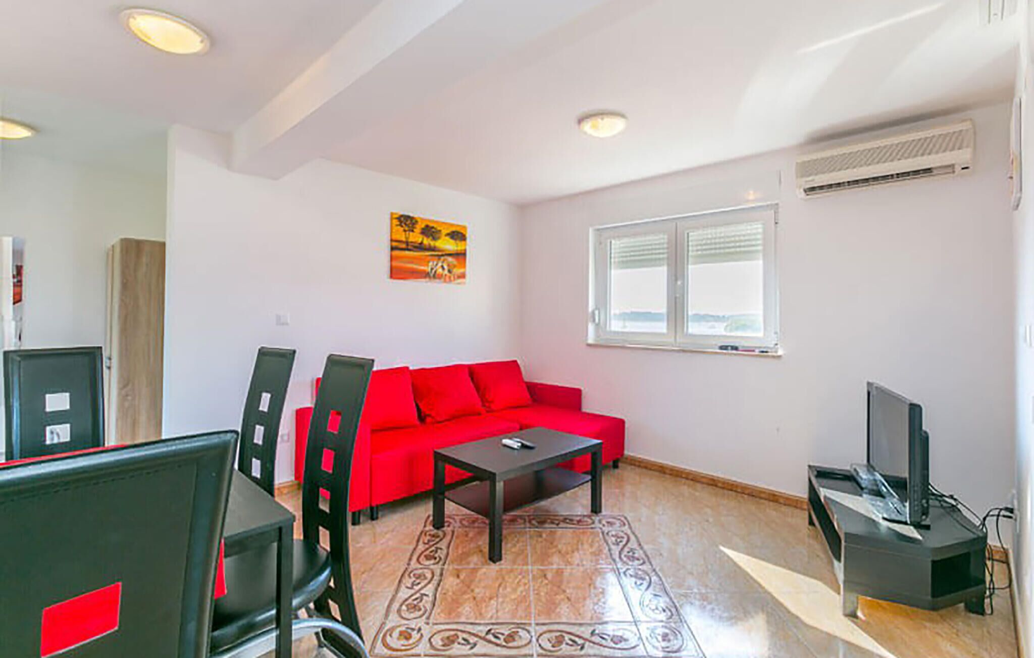 Un salon avec un canapé rouge, une petite table, un téléviseur et une fenêtre avec des stores.