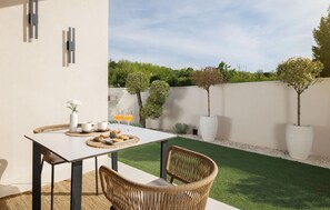 Eine Terrassengruppe im Freien mit einem Tisch für zwei Personen, umgeben von Korbstühlen, einem kleinen Garten mit Topfpflanzen und einem wolkenlosen Himmel.