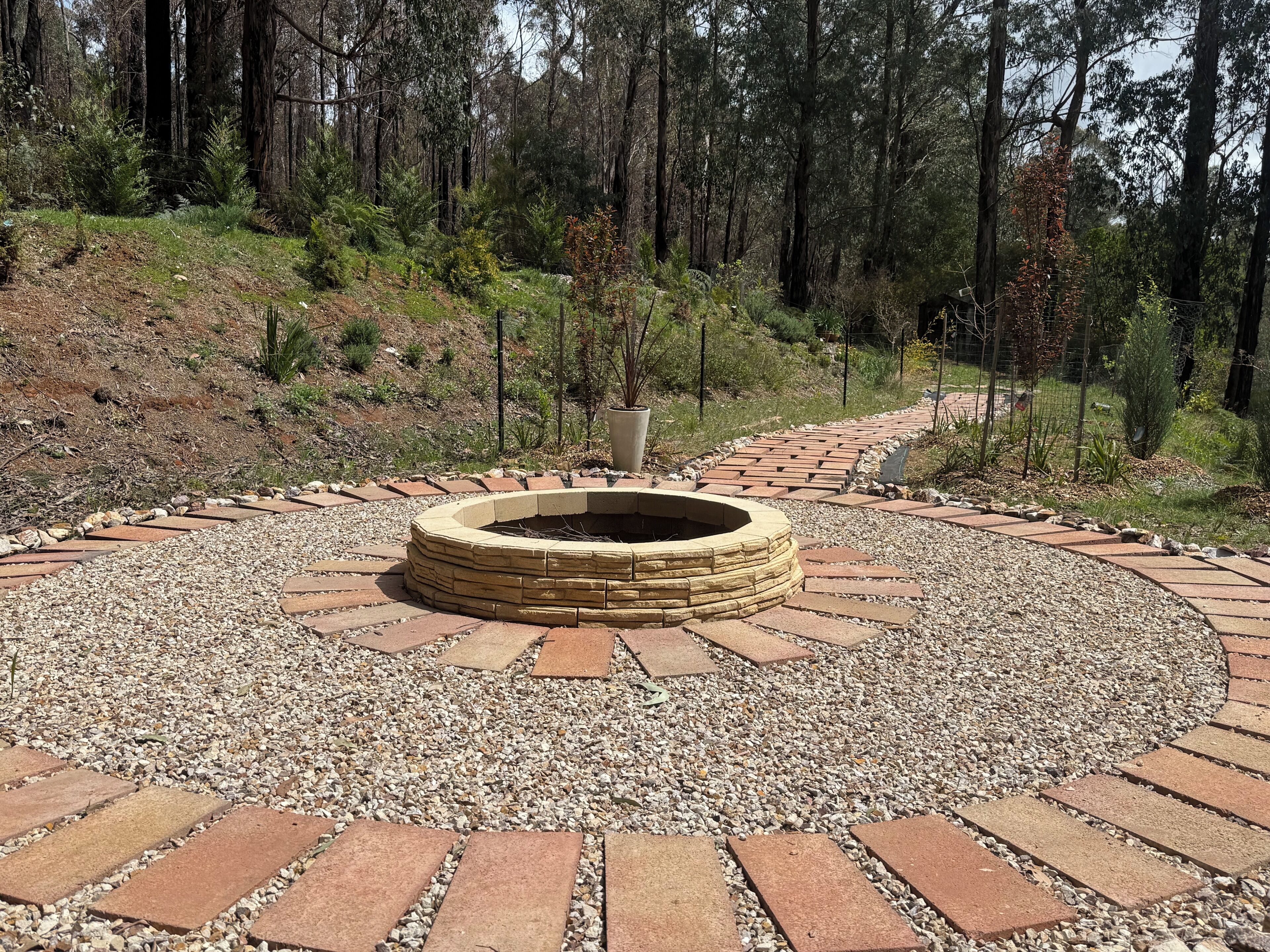 A circular stone fire pit surrounded by a gravel path, with a forested hillside in the background.