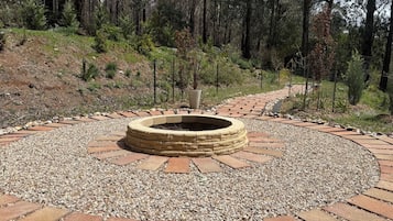 A circular stone fire pit surrounded by a gravel path, with a forested hillside in the background.