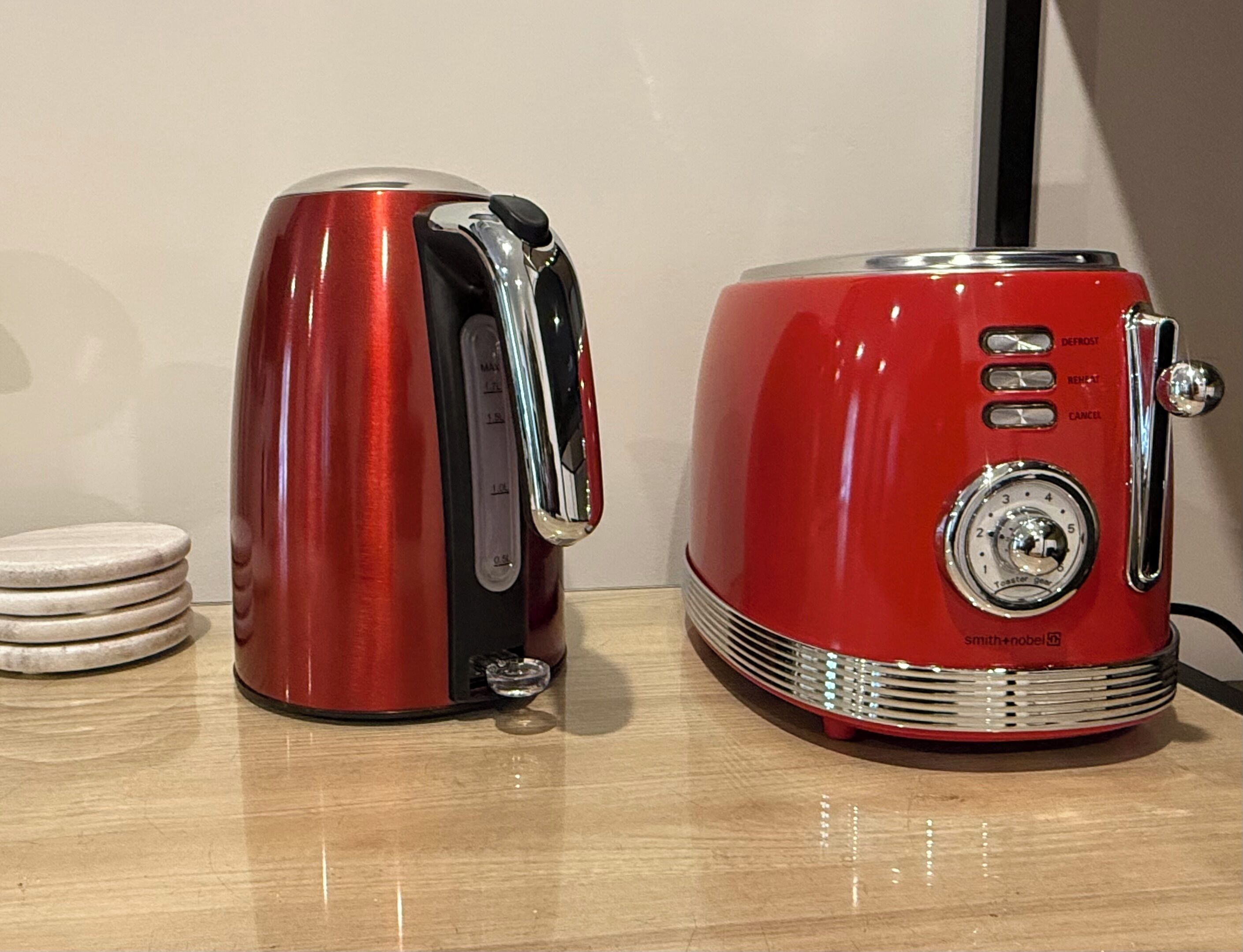 Two red appliances, a kettle and a toaster, on a wooden surface with stacked coasters.
