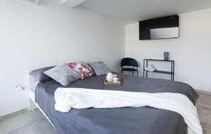 Quarto moderno com uma cama grande, uma mesa de cabeceira com abajur, uma cadeira e uma pequena mesa com toalhas.