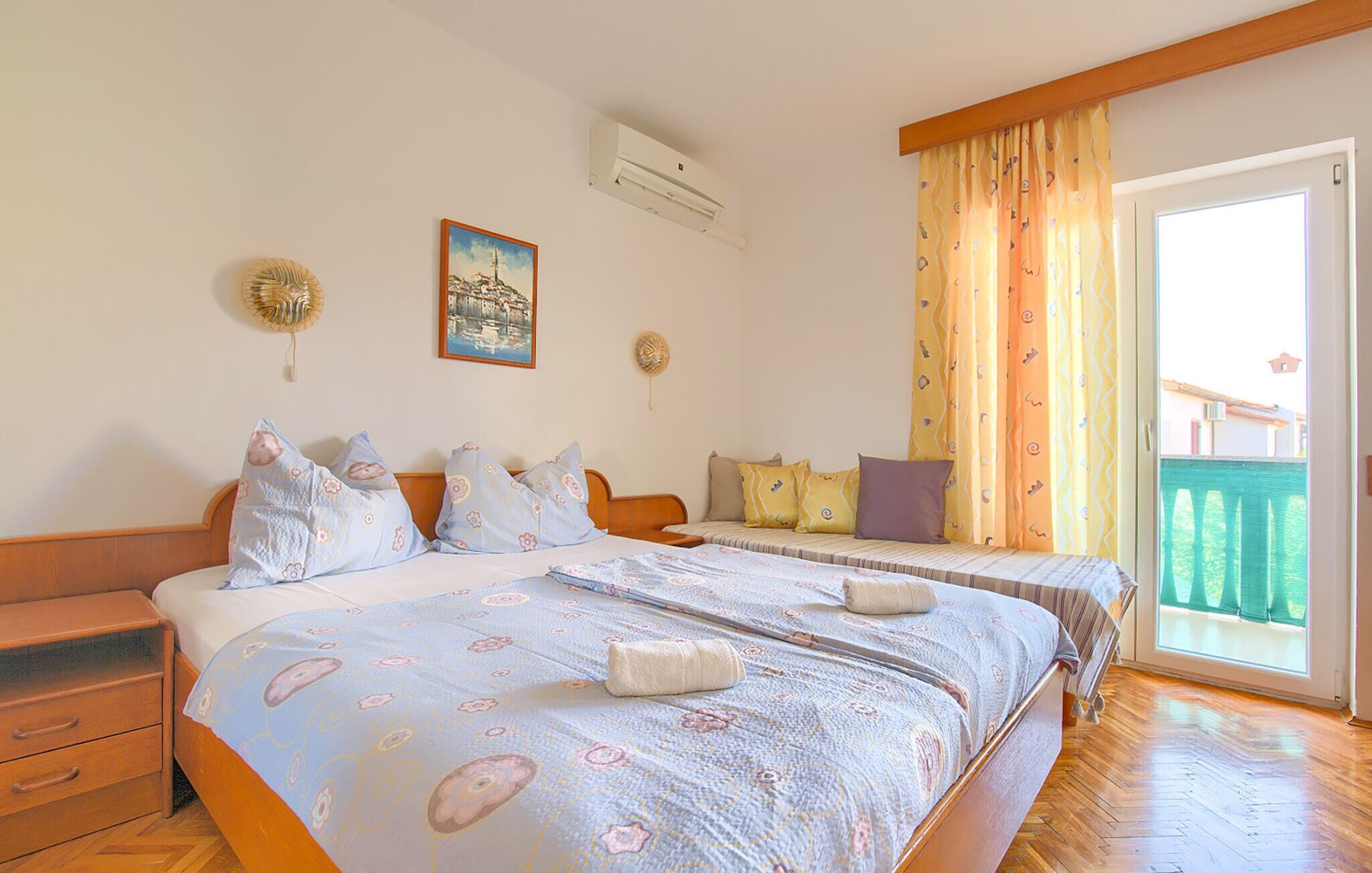 A hotel room with a large bed, a smaller bed, a wooden nightstand, a dresser, a painting on the wall, an air conditioning unit, and a view of a balcony with a railing.