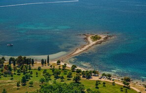 Eine Luftaufnahme einer Küstenregion mit einem Sandstrand, klarem, blauem Wasser und einer kleinen, felsigen Insel. Entlang des Ufers befinden sich Bäume und geparkte Autos.