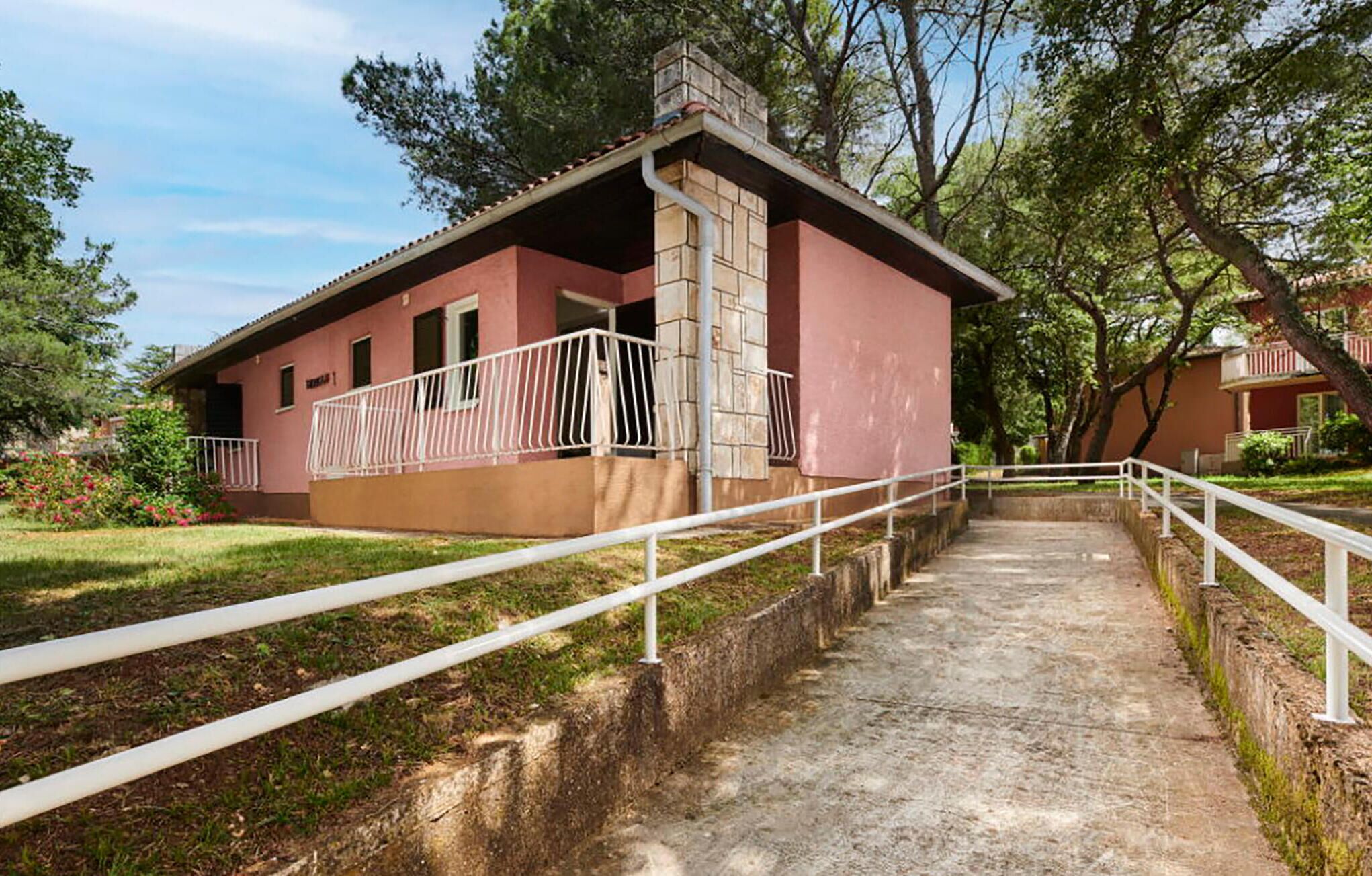 Un edificio de una planta con fachada roja, barandilla blanca a lo largo de un camino de hormigón que conduce a la entrada, y una chimenea de piedra.