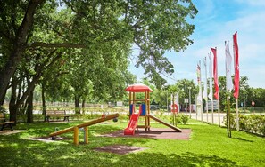A playground with a seesaw, a slide, and a climbing structure in a park with trees and flags. - Cozy apartment in Umag with outdoor swimming pool (Umag)