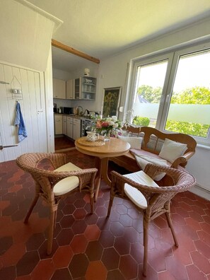 A dining area with a round table surrounded by wicker chairs, a kitchen in the background, and a view through a large window.