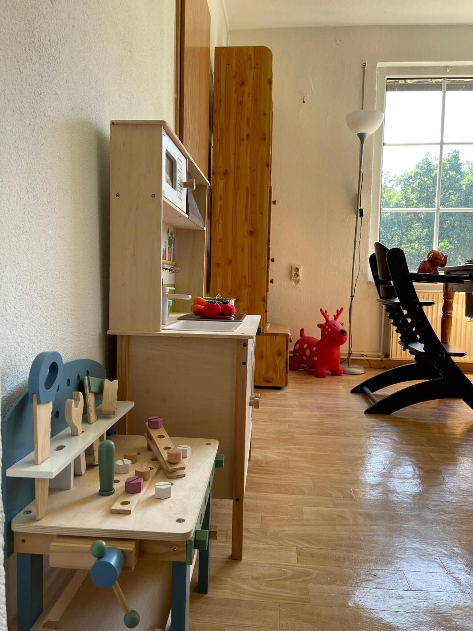 A room with a wooden table, a bookshelf, a window with a view of trees, and a red toy reindeer.