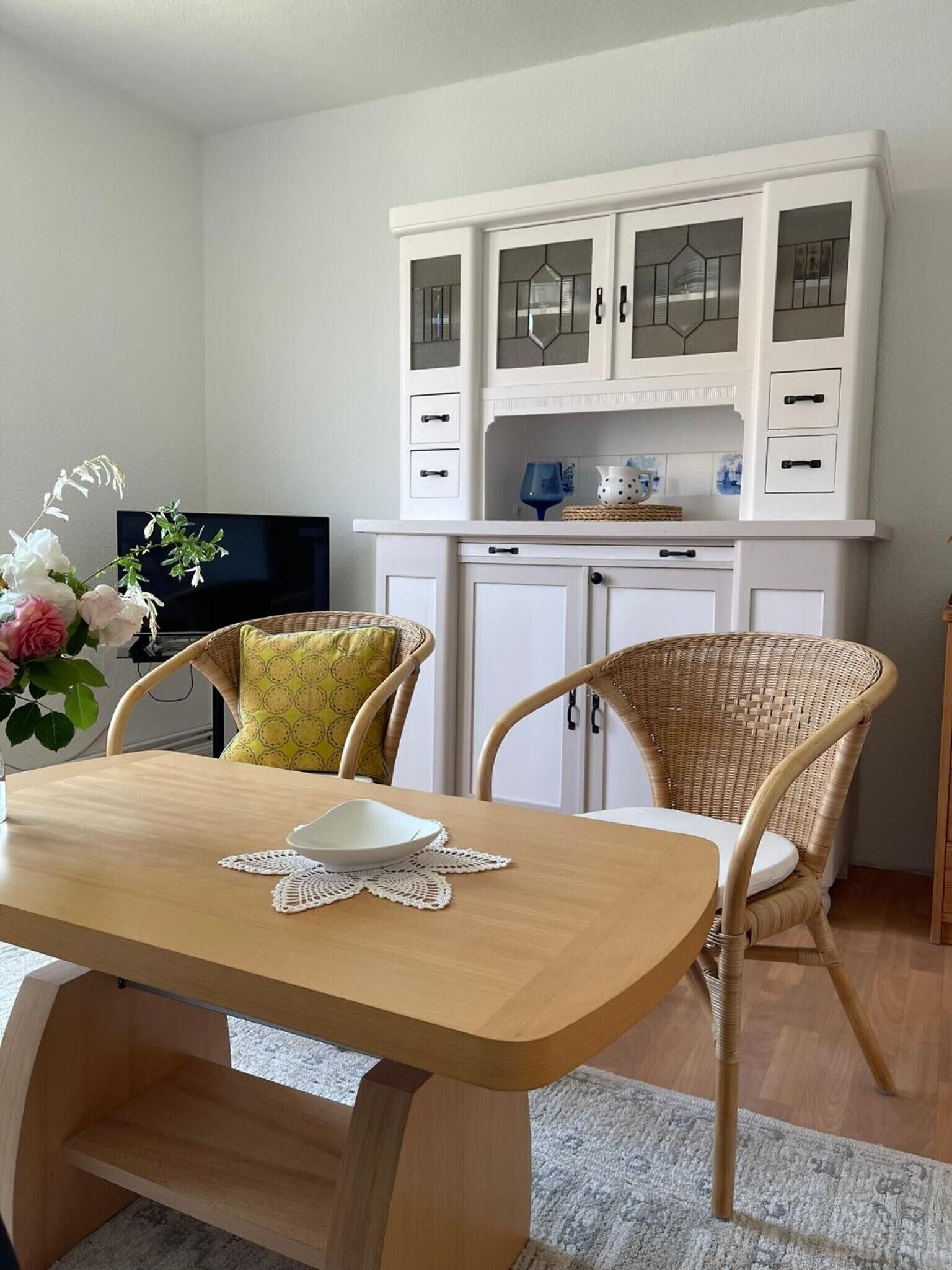 Un coin repas avec une table en bois, deux chaises en osier, un buffet blanc aux portes vitrées et un téléviseur en arrière-plan.