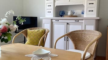 Un coin repas avec une table en bois, deux chaises en osier, un buffet blanc aux portes vitrées et un téléviseur en arrière-plan.