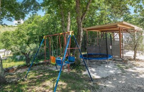 Ein Spielplatz mit Schaukeln und Trampolin, umgeben von Bäumen und einem überdachten Sitzbereich.