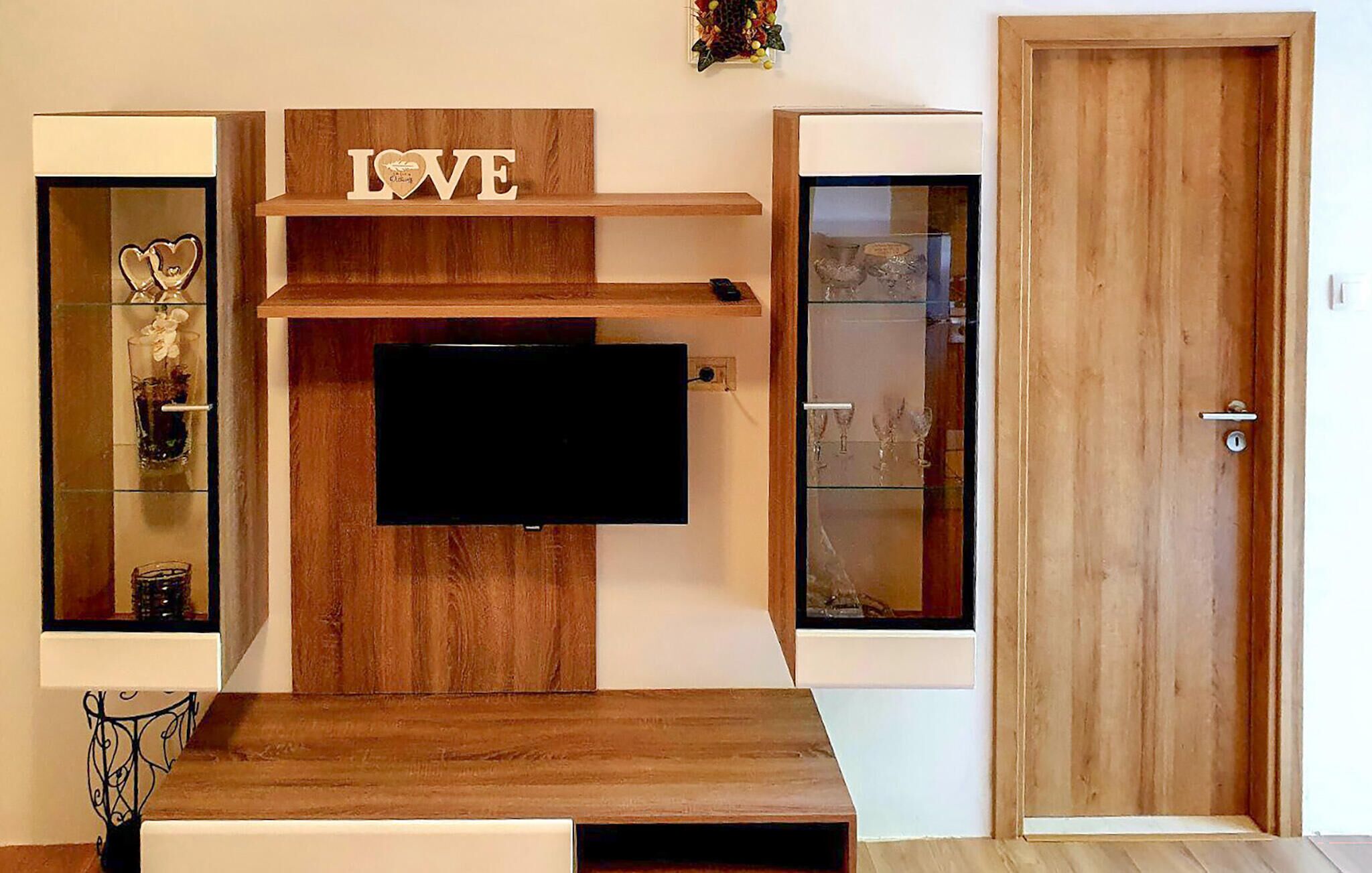 A room with a wooden wall unit above a flat-screen TV, a door to the right, and a shelf with decorative items.