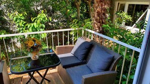 Ein Balkon mit Korbmöbeln, einem kleinen Tisch mit Blumenvase und üppiger Bepflanzung im Hintergrund.