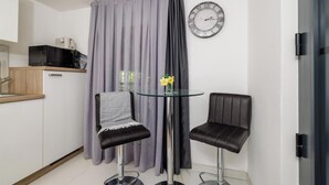 A small, modern kitchenette with a microwave, a sink, and a wooden countertop. There are two black chairs with metal bases and a glass table with a vase of yellow flowers. A large clock is on the wall, and a curtain is drawn across the window. - Beautiful apartment in Crikvenica with WiFi (Crikvenica)