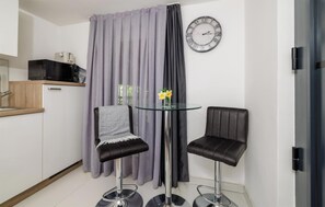 A small, modern kitchenette with a microwave, a sink, and a wooden countertop. There are two black chairs with metal bases and a glass table with a vase of yellow flowers. A large clock is on the wall, and a curtain is drawn across the window. - Beautiful apartment in Crikvenica with WiFi (Crikvenica)