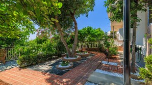 A paved garden pathway with trees and shrubs, bordered by a white fence, leading to a building entrance. - Beautiful apartment in Crikvenica with WiFi (Crikvenica)
