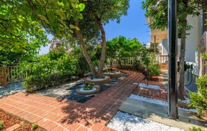 A paved garden pathway with trees and shrubs, bordered by a white fence, leading to a building entrance. - Beautiful apartment in Crikvenica with WiFi (Crikvenica)