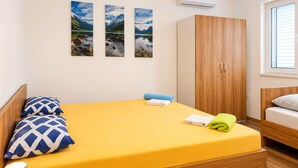 Uma cama de solteiro com colchão amarelo, duas almofadas estampadas, uma toalha azul e uma toalha verde. Há um guarda-roupa de madeira com duas portas e uma janela com persianas.