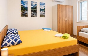 Ein Einzelbett mit gelber Matratze, zwei gemusterte Kissen, ein blaues Handtuch und ein grünes Handtuch. Ein Holzschrank mit zwei Türen und ein Fenster mit Rollläden.