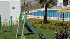 Un parque infantil con tobogán y estructura para escalar, una piscina con valla, y césped y plantas bien cuidadas.