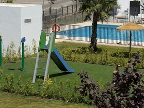 A playground with a slide and a climbing structure, a pool with a fence, and well-maintained grass and plants. - Luxury new apartment in La Cala de Mijas (Las Lagunas de Mijas)