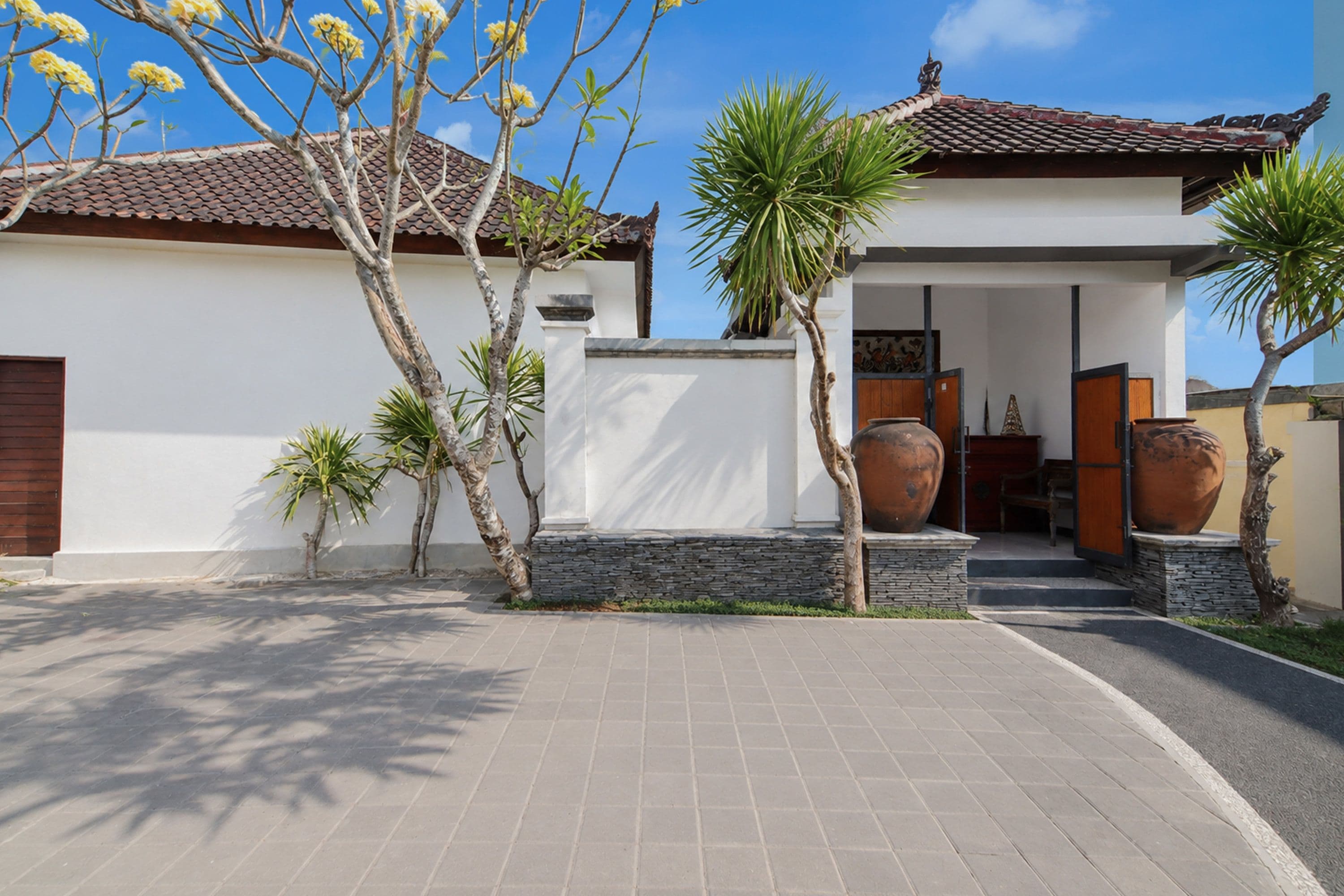 A modern house with a tiled roof, a paved driveway, and a small garden with palm trees and a large pot.