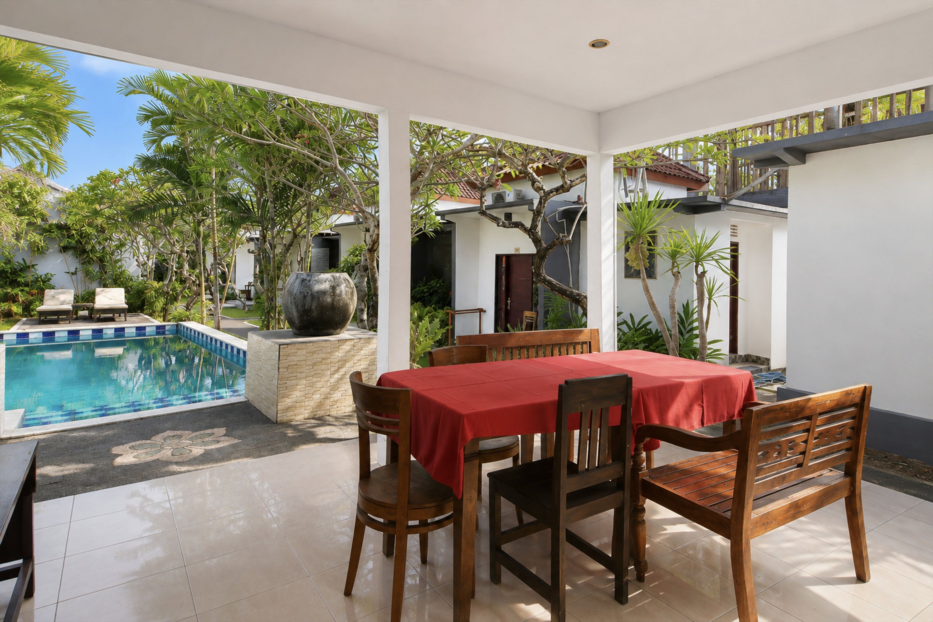 A poolside dining area with a table and chairs, a large planter, and a view of the pool and surrounding greenery.