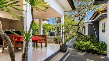A covered outdoor area with a dining table, chairs, and a bench. There are potted plants and a pathway leading to a building entrance.
