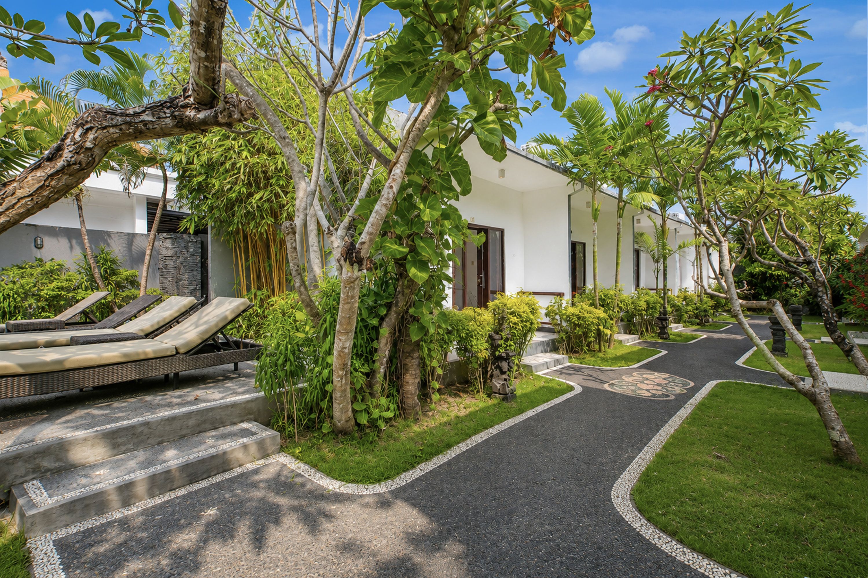 A pathway leading to a building with a modern design, surrounded by lush greenery and a seating area with lounge chairs.