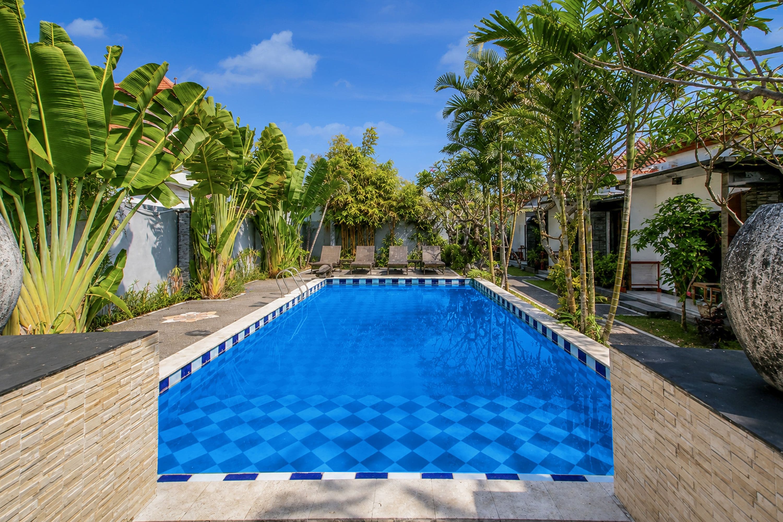 Une piscine rectangulaire aux motifs à carreaux bleus, entourée de plantes tropicales et d’un mur de pierre.