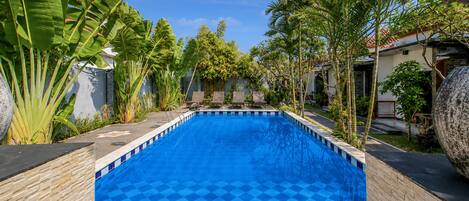 Una piscina rectangular con diseño azul a cuadros, rodeada de plantas tropicales y un muro de piedra.