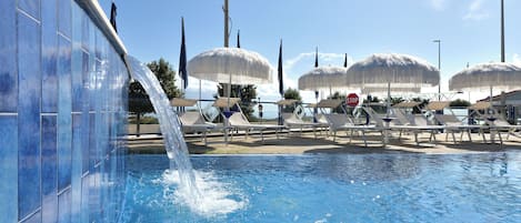 A swimming pool with a water feature, lounge chairs, umbrellas, and flags.