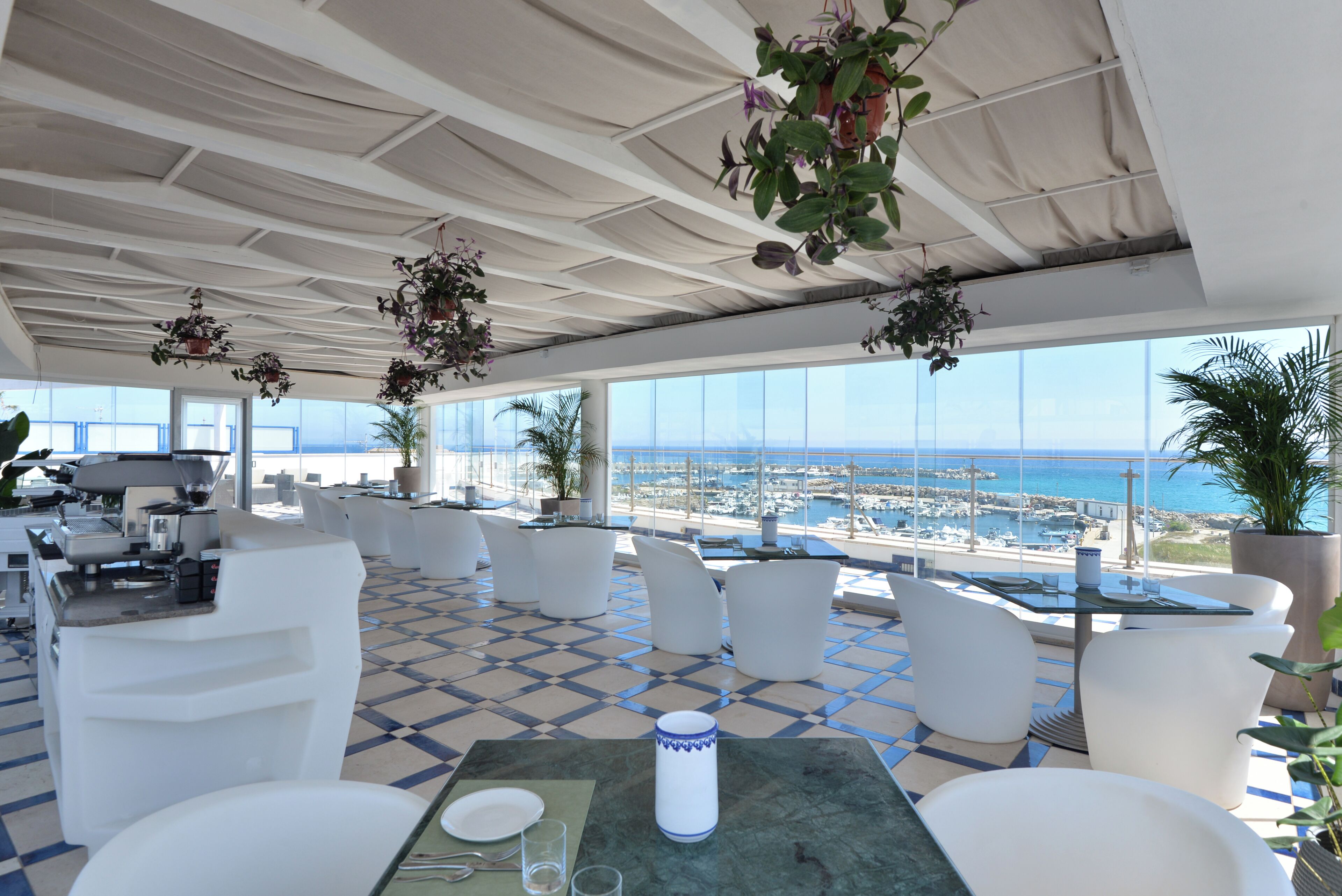 A rooftop terrace with a view of the sea, white furniture, and hanging plants.