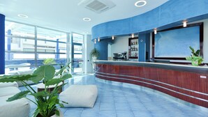 A modern reception area with a blue accent wall, a coffee machine, and a large window offering a view of the sky. - Damavi Hotel (San Foca)
