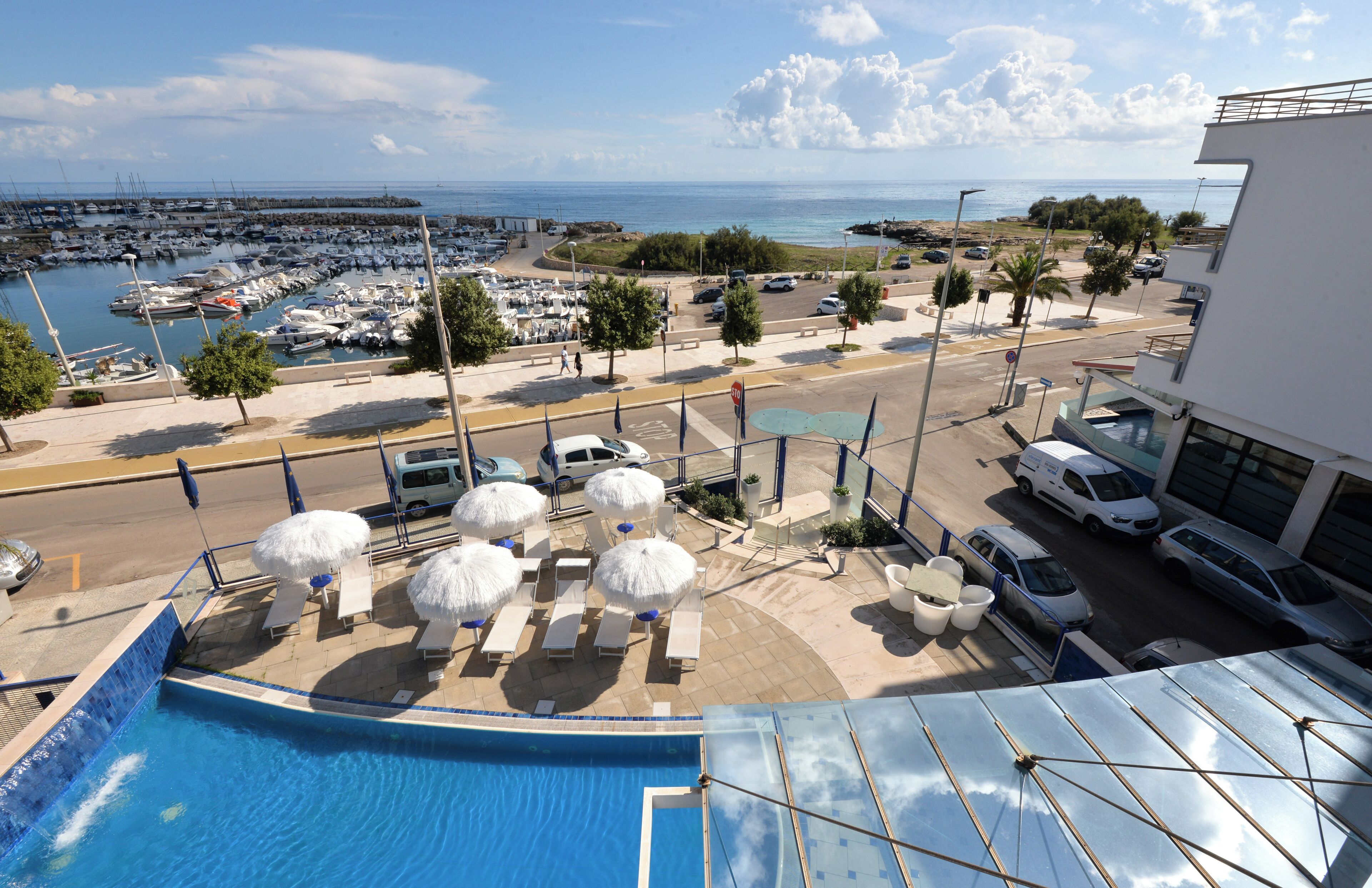 A pool area with lounge chairs and umbrellas, a marina with boats, and a view of the sea.