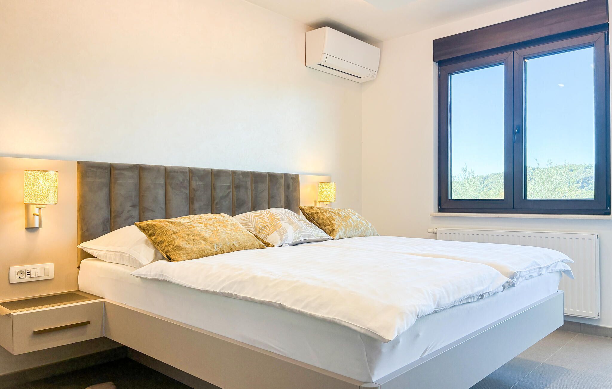 A bedroom with a bed, bedside lamps, an air conditioning unit, a window, and a radiator.